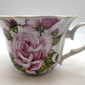 May include: A white ceramic teacup featuring a watercolor-style illustration of large pink roses, smaller rosebuds, and green leaves. The cup has a scalloped rim, a curved handle, and a smooth, glossy finish, showcasing detailed floral artwork.