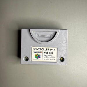May include: A grey Nintendo 64 Controller Pak memory card featuring a moulded Nintendo logo at the top. The white label displays the text CONTROLLER PAK, NINTENDO 64, NUS-004, PAT. PEND., MADE IN JAPAN, G744857, and NUS-A-PA with a colourful N64 logo.