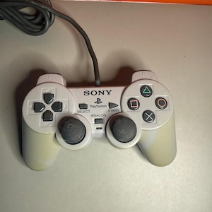 Peut inclure: Une manette de jeu filaire gris clair arborant le logo Sony PlayStation, une croix directionnelle, deux joysticks noirs, quatre boutons d'action avec des symboles géométriques, ainsi que les boutons Select, Start et Analog avec un câble gris.