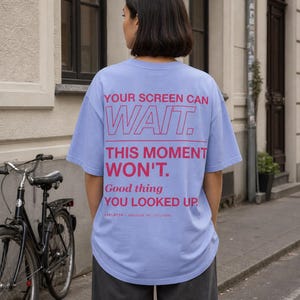 Könnte beinhalten: Ein hellviolettes T-Shirt mit kurzen Ärmeln und rotem Text auf der Rückseite: YOUR SCREEN CAN WAIT. THIS MOMENT WON'T. Good thing YOU LOOKED UP. LVPLTTR - MESSAGE 001 / COLOGNE. Das Oberteil hat einen Rundhalsausschnitt und eine lockere Passform.