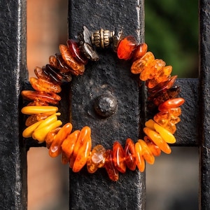 May include: A circular bracelet composed of numerous irregular, polished amber beads in shades of yellow, orange, and deep brown. The bracelet features a single metallic, ribbed cylindrical bead accent positioned at the top of the circular arrangement.