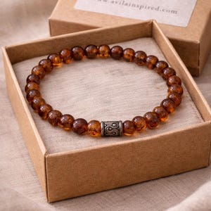 May include: A brown beaded bracelet with a silver accent piece, displayed in an open cardboard box. The box has a label reading "AVILA BY DESIGN" and a website address. The bracelet rests on a neutral-toned fabric.