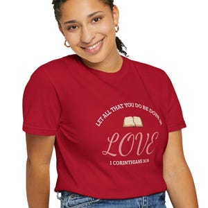 May include: A red t-shirt with the words "LET ALL THAT YOU DO BE DONE IN LOVE" and the word "LOVE" in a script font. The text also includes "1 CORINTHIANS 16:14". The shirt is paired with blue jeans.