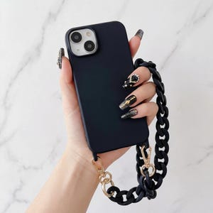 Può includere: Una custodia per telefono blu navy con una tracolla a catena nera e hardware color oro. La tracolla a catena è composta da maglie grandi e arrotondate. La custodia ha una finitura opaca e un ritaglio per la fotocamera.