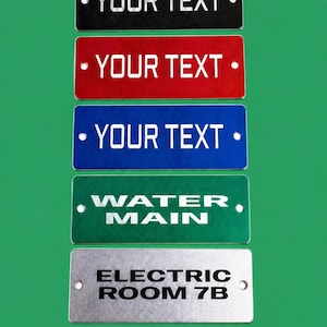 May include: Rectangular metal signs in black, red, blue, green, and silver. The top three signs read "YOUR TEXT" in white. The green sign says "WATER MAIN", and the silver sign says "ELECTRIC ROOM 7B". Each sign is 3" x 1.5".