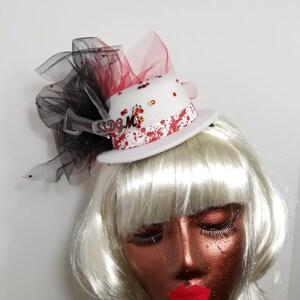 May include: White mini top hat with red and black tulle, decorated with red paint splatters, rhinestones, and a silver charm with the text "SSDGMC".