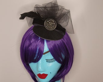 May include: A black mini hat with a black feather and a black tulle veil. The hat has a black velvet band with a silver and black decorative button.