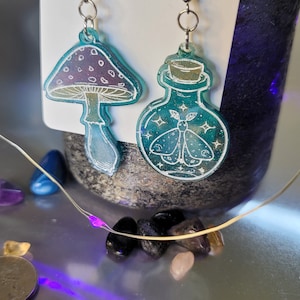 May include: A pair of earrings featuring a mushroom and a potion bottle. The mushroom is purple and white with a green stem. The potion bottle is green with a silver lid and a moth inside. Both earrings have silver hooks.