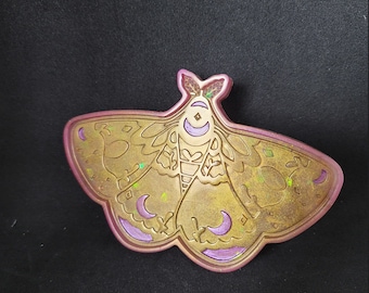 May include: A resin tray shaped like a moth with a gold and purple color scheme. The moth has a detailed design with crescent moons and floral patterns.