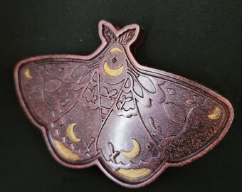May include: A dark red wooden moth pin with gold crescent moons and intricate details. The moth is in a spread wing position.