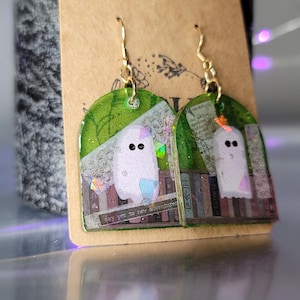 Puede incluir: Un par de pendientes verdes y dorados con un fantasma y libros en su interior. Los pendientes tienen forma de rectángulo con esquinas redondeadas. El texto en los libros dice "Di sí a nuevas aventuras".