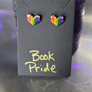 May include: A pair of rainbow pixel heart stud earrings on a black card with the text "Book Pride" written in yellow.