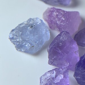 May include: A collection of raw, unpolished gemstones in various shades of purple and light blue. The stones feature irregular, jagged edges and translucent surfaces with crystalline textures, scattered across a plain, light-colored surface.