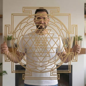 May include: A golden-coloured, geometric wall art piece with a complex, layered design, including a central triangle and circular patterns. The art is held by a person in a white t-shirt. Suitable for home decor.