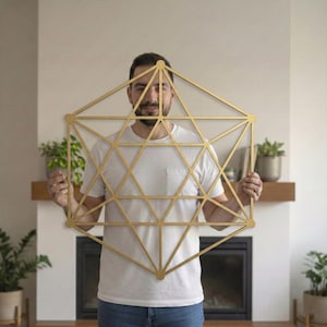 May include: A gold-colored geometric wall art piece. The artwork features a complex pattern of interconnected lines and shapes, forming a star-like design. The piece is held up in front of a person. The artwork is approximately 30 inches in height.