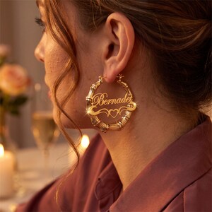Puede incluir: Un pendiente de aro dorado estilo bambú con una placa personalizada en letra cursiva que dice Bernadet, acentuado con un recorte decorativo en forma de corazón y adornos en espiral integrados en el centro del diseño circular.