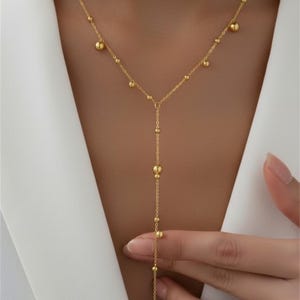 Op de afbeelding: Een goudkleurige ketting met een Y-vormig ontwerp. De ketting heeft kleine, ronde kralen die gelijkmatig over de ketting zijn verdeeld en een langere verticale druppel met grotere, decoratieve kralen. De ketting wordt door een persoon vastgehouden.