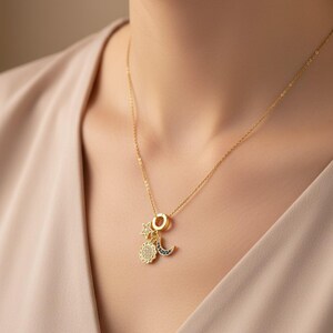 Puede incluir: Un collar dorado con colgantes celestiales. El collar presenta un colgante de sol, luna y estrella. Los colgantes de sol y luna están adornados con pequeñas piedras brillantes. El collar lo lleva una persona.