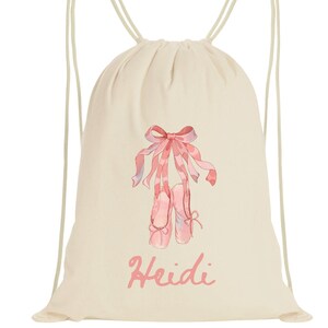 May include: A cream-colored drawstring backpack featuring a watercolor design of pink ballet shoes with a matching bow and ribbons. The name "Heidi" is written in pink script below the ballet shoes. The bag has cream-colored drawstrings.
