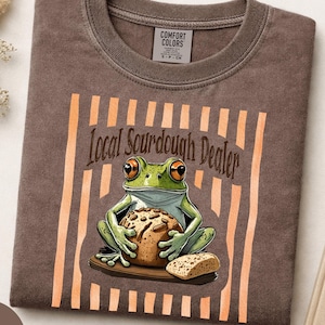 Op de afbeelding: Een bruin t-shirt met een afbeelding van een groene kikker die een brood vasthoudt. Boven de kikker staat de tekst Local Sourdough Dealer. Het ontwerp is geplaatst tegen een achtergrond van verticale oranje strepen. Het shirt heeft een Comfort Colors label.