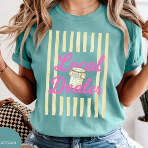Op de afbeelding: Een zeegroen t-shirt met korte mouwen en een grafische print van verticale lichtgele strepen. In het midden staat een tekening van een glazen pot met een deksel en de roze tekst Local Dealer in een cursief lettertype.