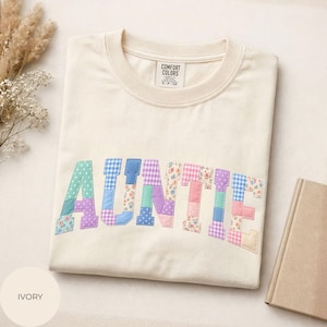 Könnte beinhalten: Ein elfenbeinfarbenes T-Shirt mit dem Wort AUNTIE in großen Blockbuchstaben. Jeder Buchstabe besteht aus verschiedenen Stoffflicken in Pastellfarben, darunter Blumenmuster, Vichy-Karos und Punkte. Das Shirt hat ein Comfort Colors Etikett.