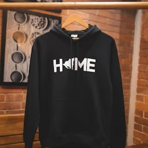 Può includere: Felpa con cappuccio nera con la scritta bianca "HOME". La "O" in "HOME" è sostituita da un disegno bianco di uno stato. La felpa ha una tasca frontale e un cappuccio con coulisse. È appesa a una gruccia di legno.