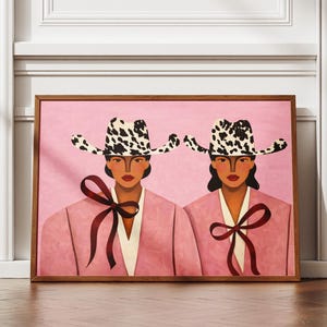 May include: A framed rectangular wall art print featuring two figures wearing cow-print cowboy hats and pink jackets with dark red ribbon bows tied at the neck. The figures are set against a solid pink background with a smooth, matte finish.