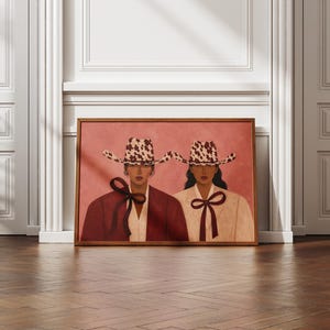 May include: A framed rectangular wall art print featuring two individuals wearing matching cow-print cowboy hats and collared shirts with dark ribbon ties. One individual wears a deep red jacket, while the other wears a light beige jacket against a pink background.