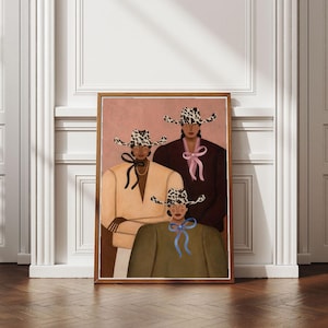 May include: A framed art print depicting three figures wearing cow-print cowboy hats. The figures are dressed in solid-colored tops in beige, dark brown, and olive green, each accented with a distinctively colored ribbon tie in black, pink, or blue.