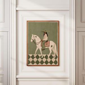 May include: A framed wall art print featuring a person wearing a wide-brimmed hat and light-colored clothing riding a white horse. The horse has dark spots and a pink saddle blanket. The background is olive green with a checkered floor pattern.
