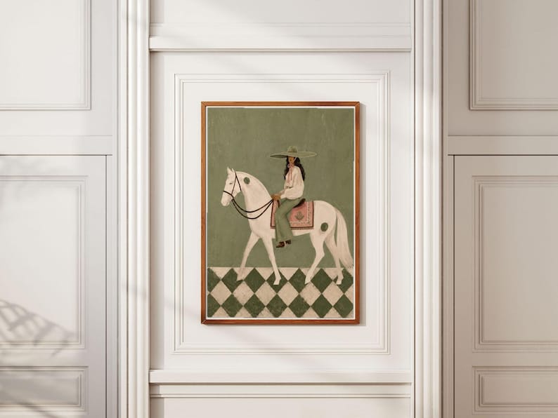 May include: A framed wall art print featuring a person wearing a wide-brimmed hat and light-colored clothing riding a white horse. The horse has dark spots and a pink saddle blanket. The background is olive green with a checkered floor pattern.