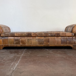May include: A long, low-profile bench upholstered in a patchwork of brown leather squares. The piece features two rounded, rolled arms on each end and a tufted, rectangular seat cushion, showcasing varied shades of tan, chestnut, and dark brown leather.