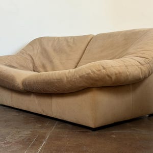 May include: A low-profile, two-seater sofa upholstered in tan suede. The piece features a continuous, rounded backrest that flows into wide, padded armrests, creating a unified, soft silhouette with a rectangular base and visible seams.