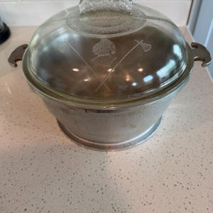 Puede incluir: Una fuente para horno plateada vintage con tapa de cristal transparente. La tapa presenta un diseño grabado de palos de lacrosse cruzados y un casco. La fuente tiene un exterior texturizado y un asa de cristal transparente.