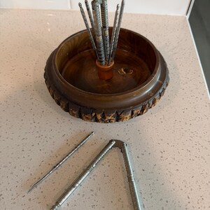 May include: A wooden nut bowl with a decorative edge, holding several silver nut picks. A silver nutcracker and two silver picks are placed in front of the bowl. The bowl and tools are on a light-colored surface.