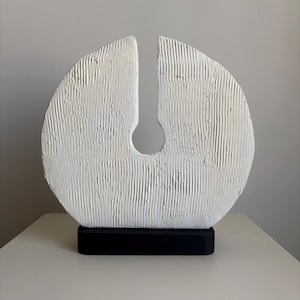 Op de afbeelding: Een witte cirkelvormige sculptuur met een getextureerd, geribbeld oppervlak en een centrale verticale uitsparing die eindigt in een rond gat. De sculptuur is gemonteerd op een stevige, rechthoekige zwarte voet die contrasteert met de witte vorm.