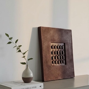 Puede incluir: Una pieza de arte mural cuadrada marrón texturizada con una cuadrícula de recortes rectangulares. Un pequeño jarrón de dos tonos con una ramita de vegetación y un libro titulado "RULKENTER OMED X ASTAR" están en un estante.