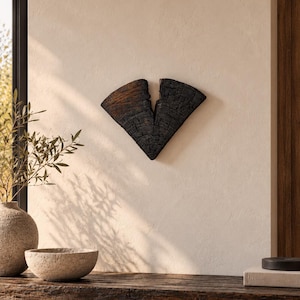 Può includere: Un oggetto decorativo da parete composto da due pezzi di legno carbonizzato a forma di V con una visibile consistenza screpolata. Il legno presenta toni scuri profondi e una finitura superficiale bruciata e ruvida, disposti in una forma divisa.
