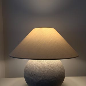 Puede incluir: Una lámpara de mesa moderna con una base esférica texturizada y una pantalla cónica. La base es de color gris claro y la pantalla es de color beige. La lámpara está encendida, emitiendo un brillo cálido.