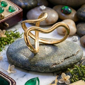 Peut inclure: Une bague dorée au design fluide unique repose sur une pierre grise. L'image présente également des émeraudes, des opales et des diamants, ainsi qu'un petit flacon de parfum et une plaque dorée.