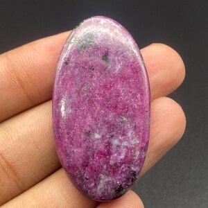 Pode incluir: Uma pedra preciosa em formato de cabochão oval com uma cor base magenta e rosa vibrante, apresentando inclusões minerais pretas e brancas espalhadas por toda a superfície polida, com uma textura lisa e arredondada e um acabamento brilhante.