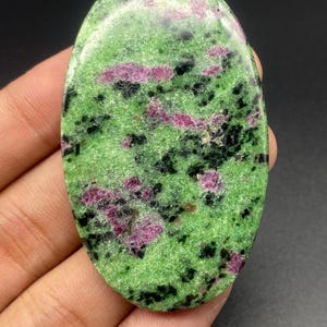 May include: An oval-shaped polished gemstone featuring a vibrant green base color with scattered patches of deep magenta and dark black inclusions throughout the surface. The stone has a smooth, glossy finish and a distinct, mottled appearance.
