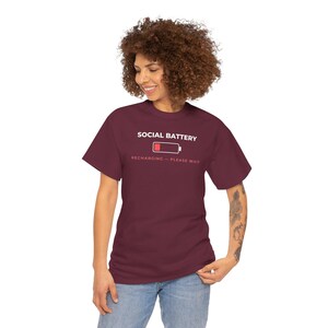 May include: A maroon short-sleeved t-shirt featuring the white text SOCIAL BATTERY above a graphic of a low-charge battery icon with a red segment, followed by the text RECHARGING - PLEASE WAIT in a smaller, lighter red font centered on the chest.