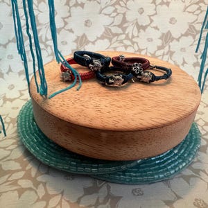 以下が含まれることがあります： 赤、ネイビー、黒の手作りのブレスレットのコレクション。それぞれにカメのチャームが付いています。ブレスレットは、ターコイズ色のビーズコースターの上に置かれた木製のプラットフォームに展示されています。背景にはターコイズ色の糸が垂れ下がっています。