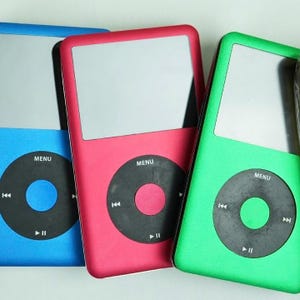 May include: Three digital music players in blue, red, and green. Each device features a screen, a circular control pad with a menu button, and playback controls. The players are arranged side by side, highlighting their bright colours.