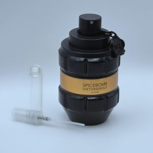 Könnte beinhalten: Schwarze und goldfarbene Spicebomb Viktor & Rolf Extreme Duftflasche mit einem kleinen, klaren Glasfläschchen und einem transparenten Kunststoffzerstäuber. Die Flasche hat ein granatenähnliches Design mit dem Markennamen auf einem goldenen Band.