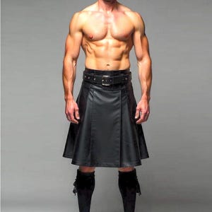 Op de afbeelding: Een zwarte leren kilt met een brede riem en gesp. De kilt wordt gecombineerd met zwarte kniehoge sokken en zwarte schoenen. De afbeelding is tegen een grijze achtergrond.