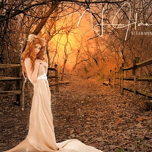 May include: A woman with long, red hair stands in a forest setting. She is wearing a long, flowing white dress and is looking down. The background is a blurred image of trees and a path. The image is in a sepia tone.