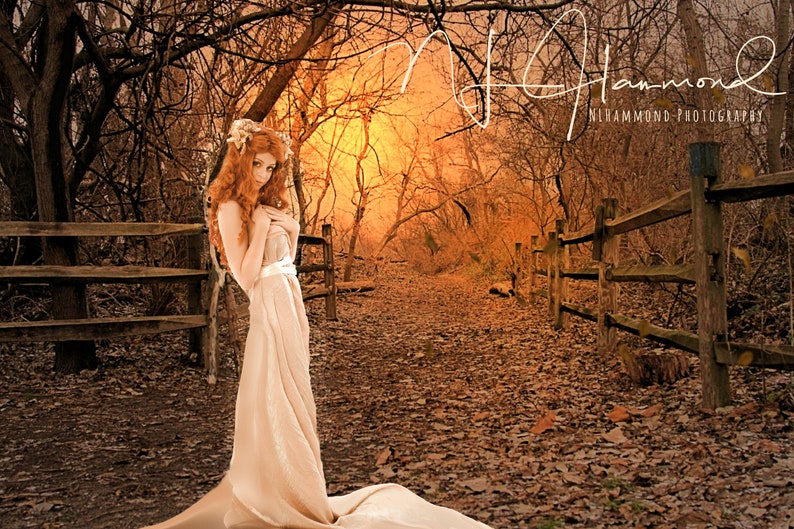 May include: A woman with long, red hair stands in a forest setting. She is wearing a long, flowing white dress and is looking down. The background is a blurred image of trees and a path. The image is in a sepia tone.